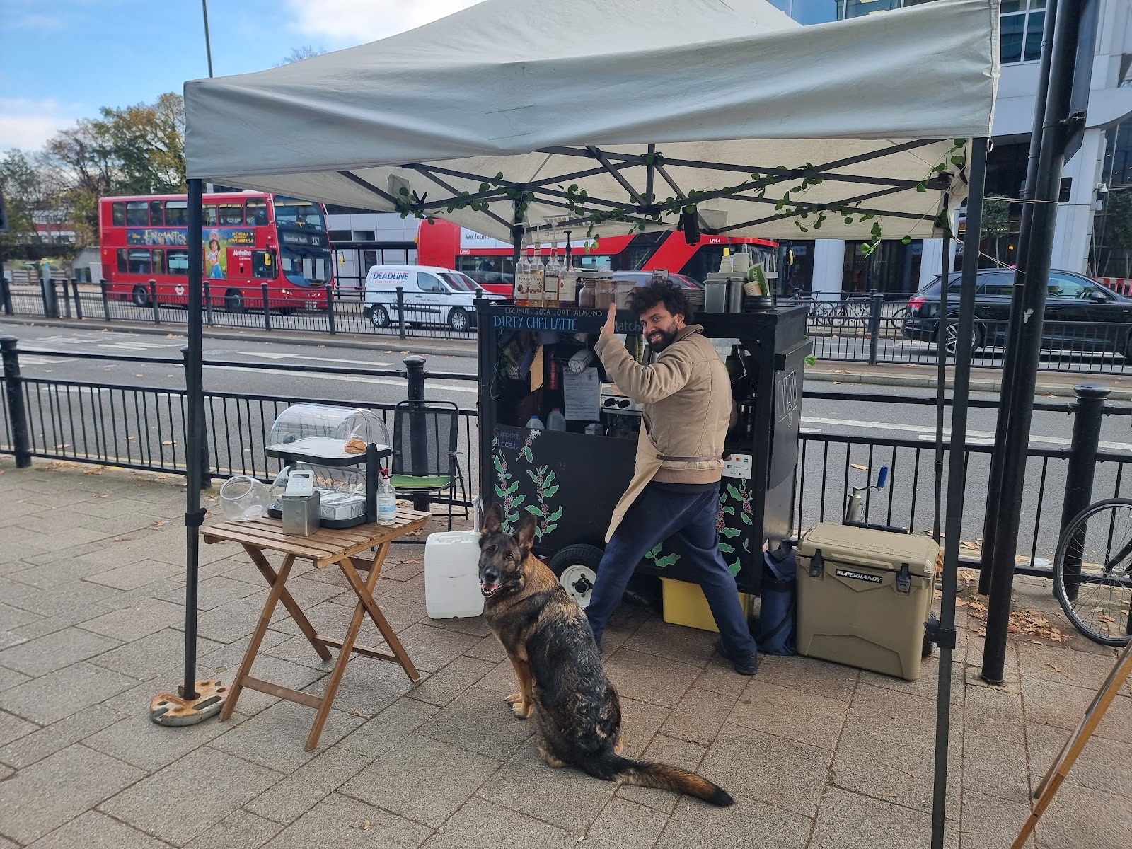 The barista making coffee at the cart, a dog resting in front and a red bus passing behind