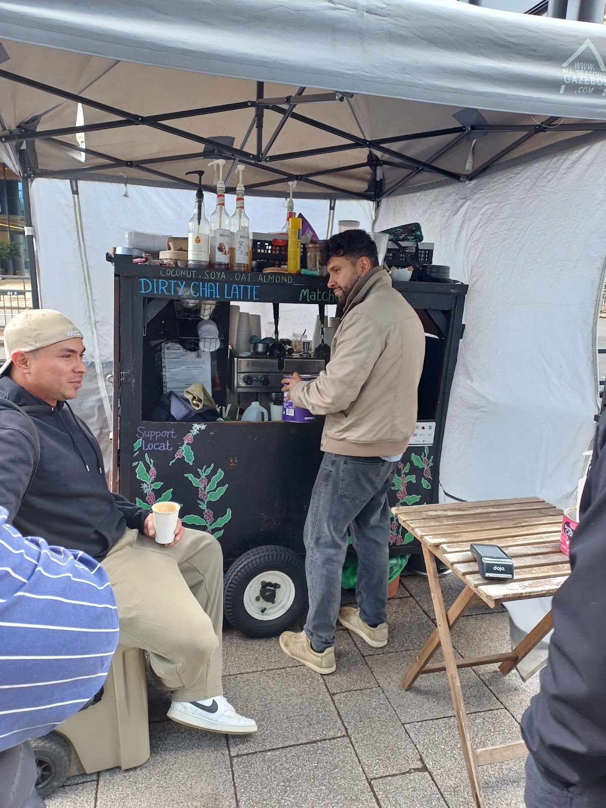 Customers at the Cafetto coffee cart under the gazebo, one seated with a cup and one waiting at the machine